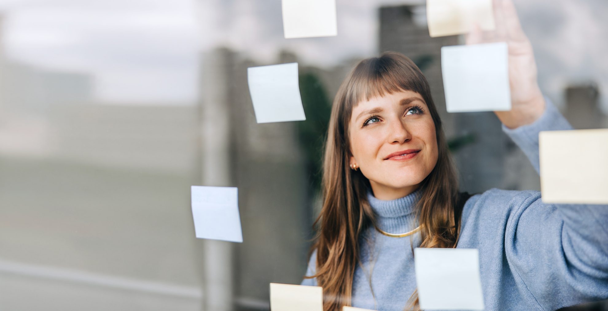 2176610625 Young female entrepreneur sticking adhesive notes to a glass wall in a modern office. Happy young businesswoman laying out her ideas while working on a new creative strategy.