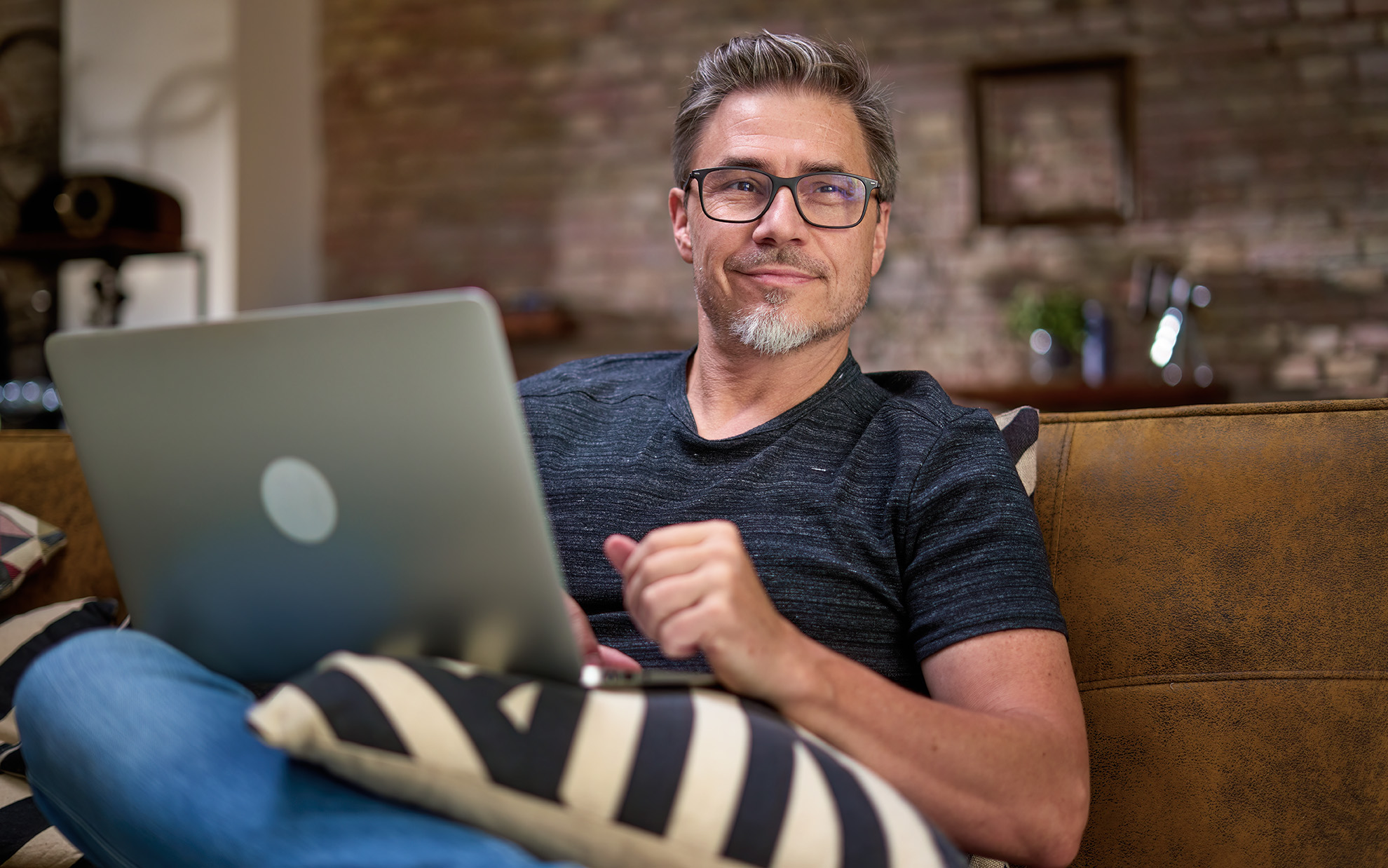 Older man working online with laptop computer at home sitting on couch in living room. Home office, browsing internet. Portrait of happy, mature age, middle age, mid adult man in 50s, smiling.