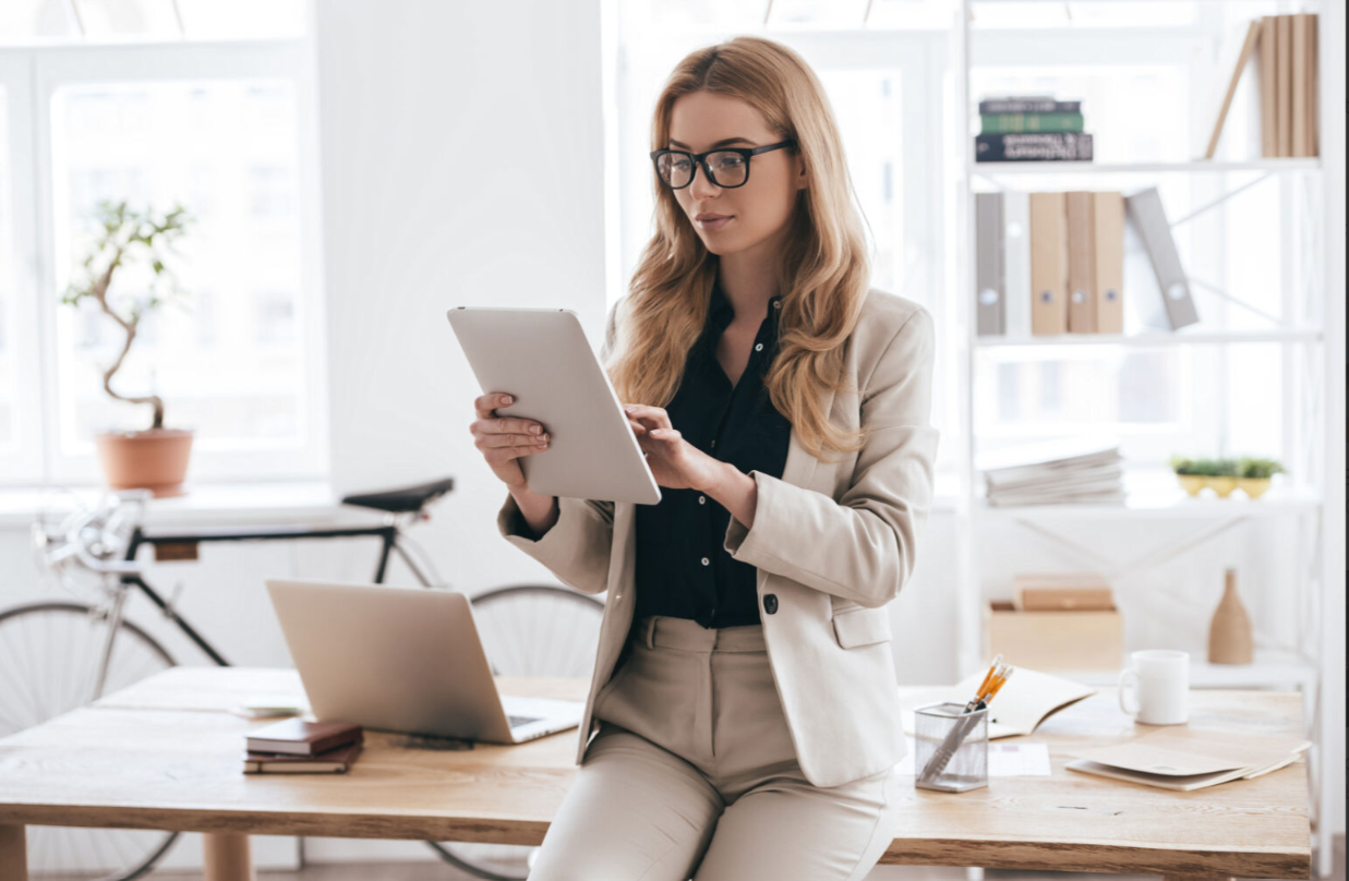 Mujer profesional trabajando desde un espacio luminoso, vestida con traje y usando una tablet. Representa el liderazgo moderno y la gestión efectiva en entornos de teletrabajo.