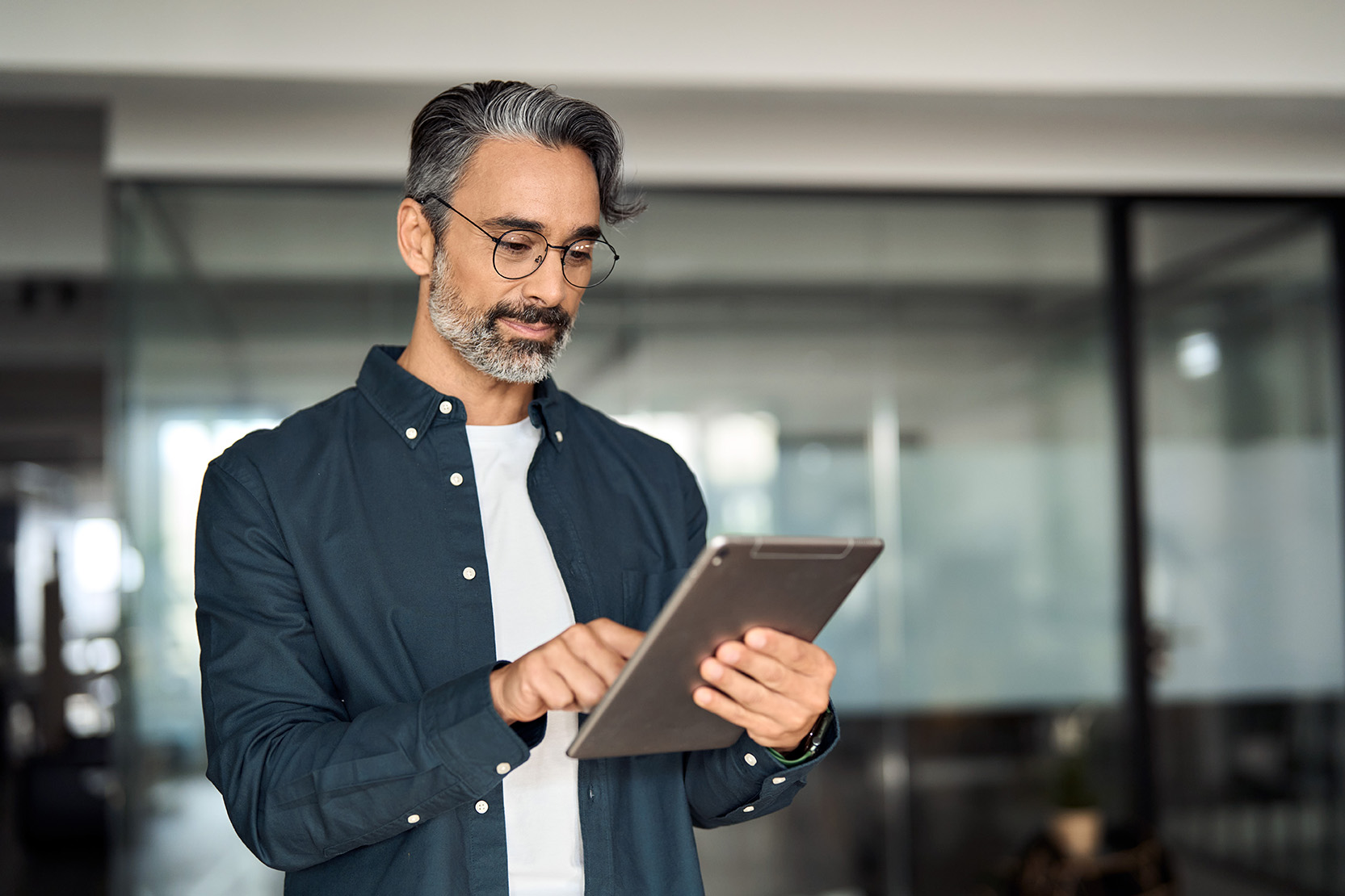 Busy middle aged business man wearing shirt standing in corporate office using digital tablet. Mature older businessman professional executive manager investor working on fintech tab computer device.