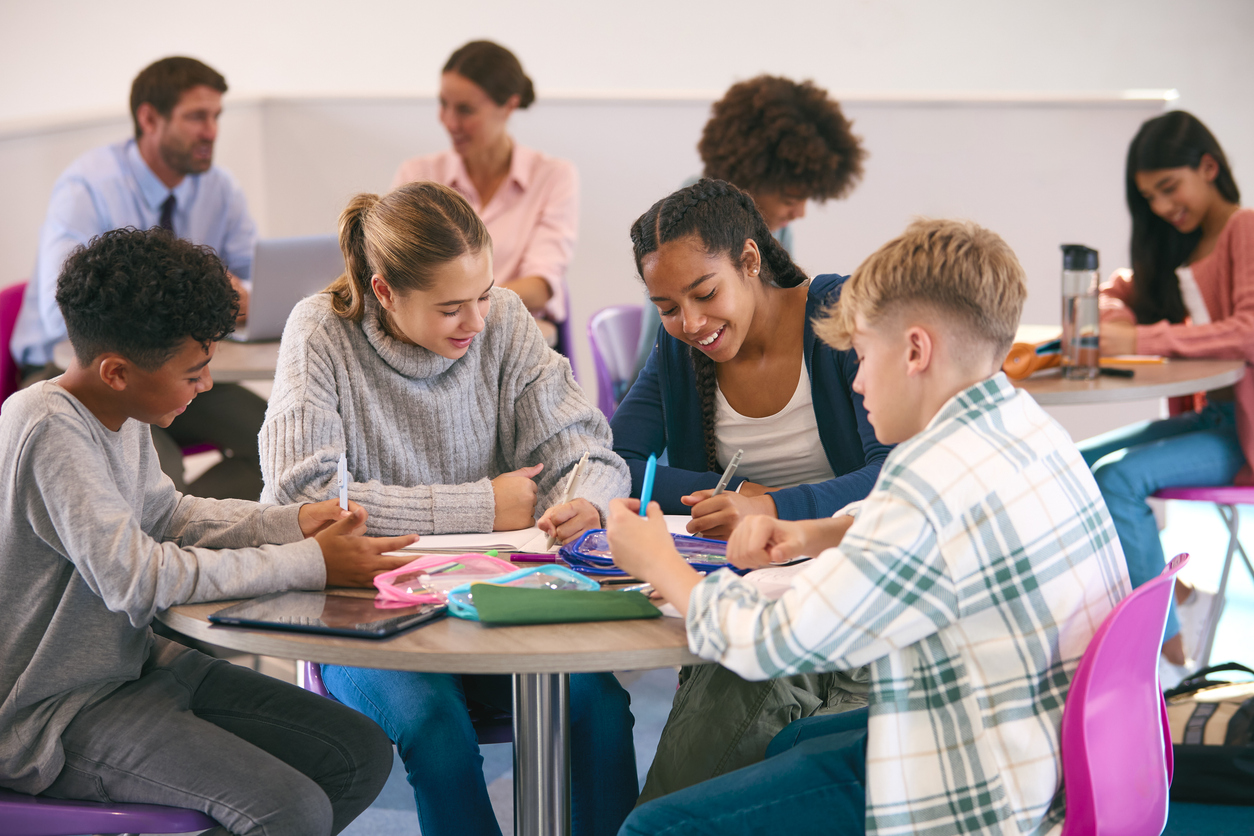 Secondary Or High School Students Collaborating In Study Area With Teachers In Background