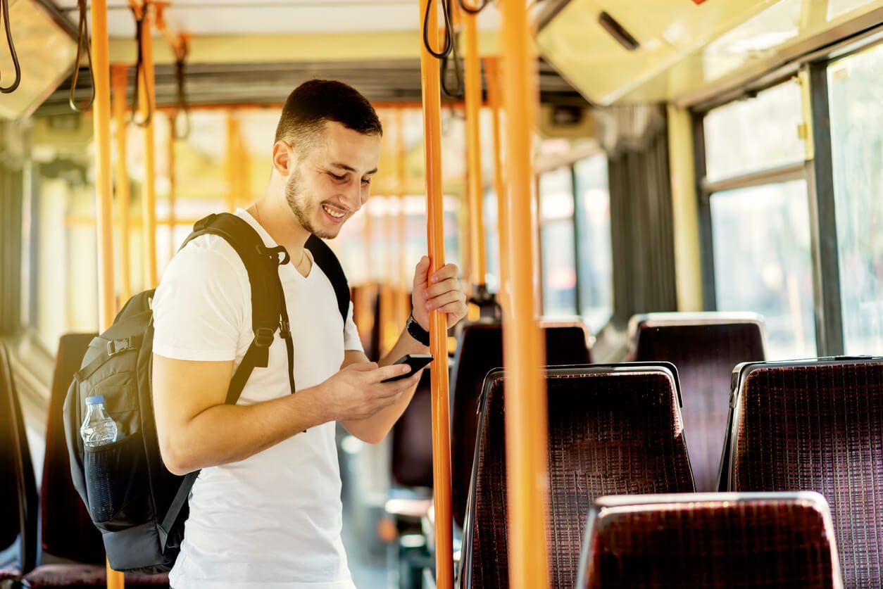 um estudante de pé no ônibus, de mochila nas costas, segurando a barra com uma das mãos e o celular com a outra