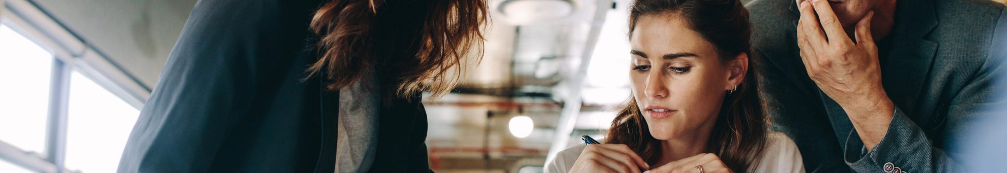 Mulheres profissionais analisando documentos sobre uma mesa em uma reunião de trabalho, enquanto colaboram com um colega em um escritório moderno.