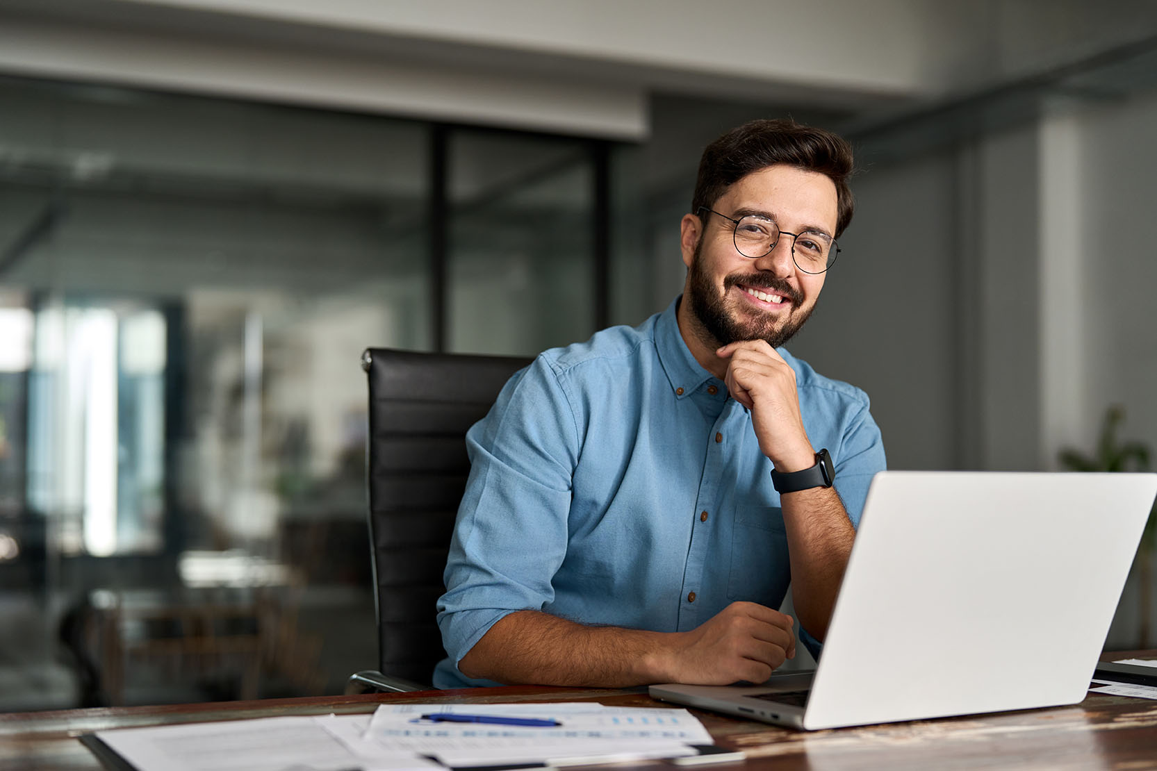 Portrait of happy professional business man company employee, young smiling male businessman entrepreneur or worker looking at camera using laptop computer technology working at corporate office desk.