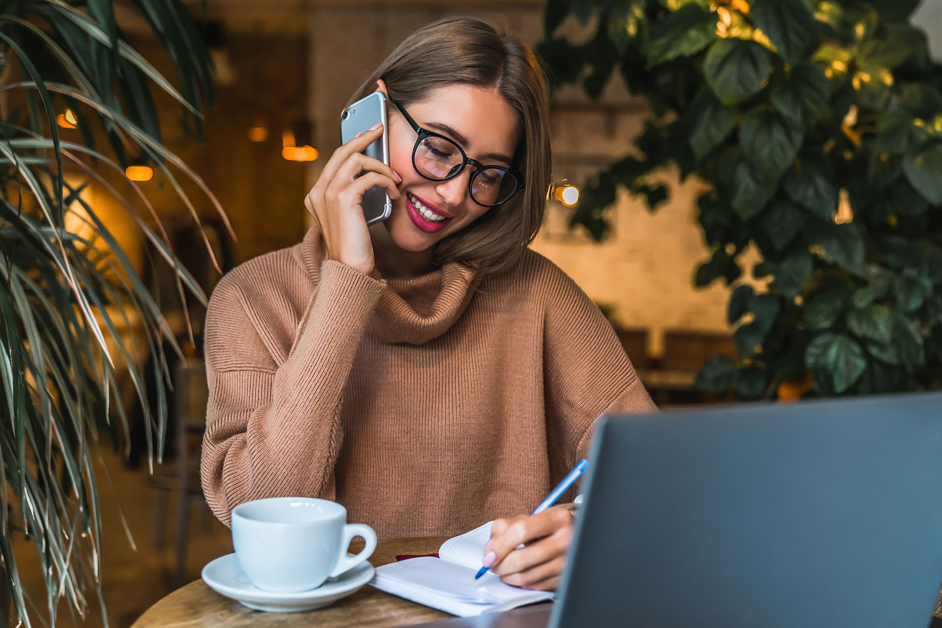 Young woman sitting in a restaurant busy working and talking on mobile phone. Female freelancer working on laptop computer making notes at modern loft cafe . Entrepreneur have work call while break